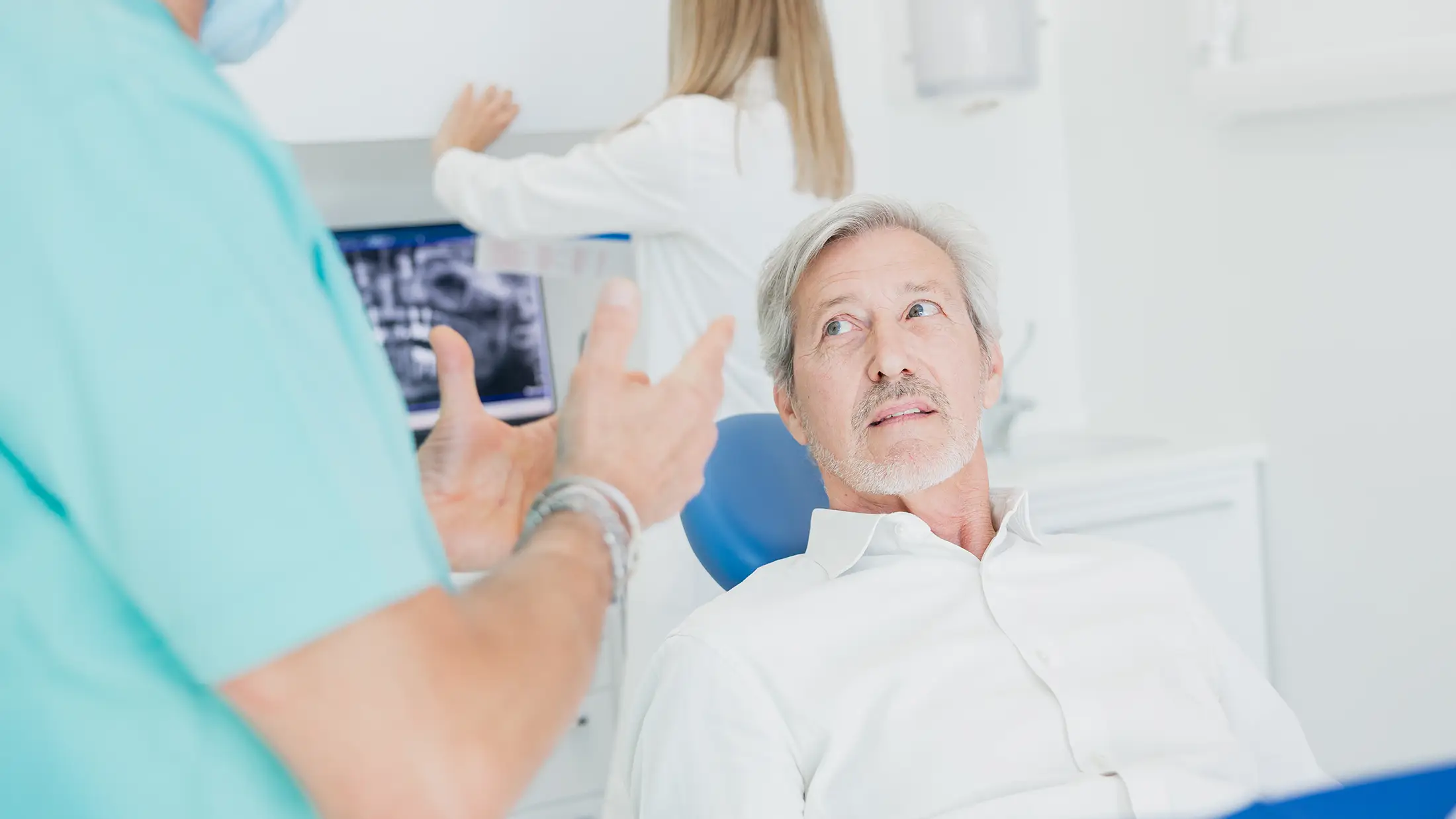 Un paziente anziano siede sulla poltrona odontoiatrica mentre dialoga con un dentista che gesticola durante la spiegazione di un trattamento preventivo nello Studio Confident.